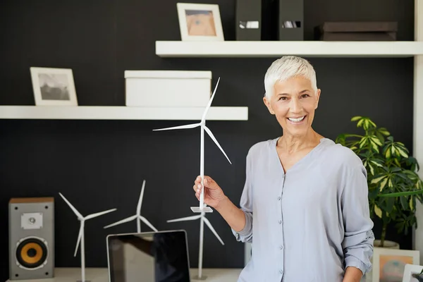 Hermosa mujer mayor caucásica sonriente de pie en la oficina y sosteniendo el modelo de molino de viento. Concepto de creación de empresas . — Foto de Stock