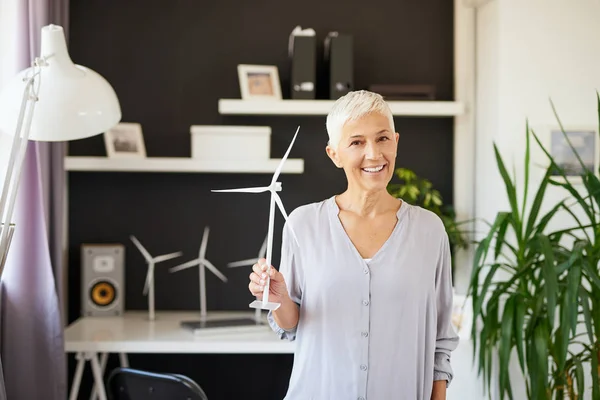 Hermosa mujer mayor caucásica sonriente de pie en la oficina y sosteniendo el modelo de molino de viento. Concepto de creación de empresas . — Foto de Stock