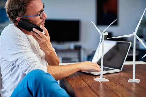 Beau ingénieur écologiste caucasien assis à la maison, parlant au téléphone du projet et tenant le modèle de moulin à vent. Sur le bureau sont des modèles de moulin à vent. Concept de développement durable . — Photo