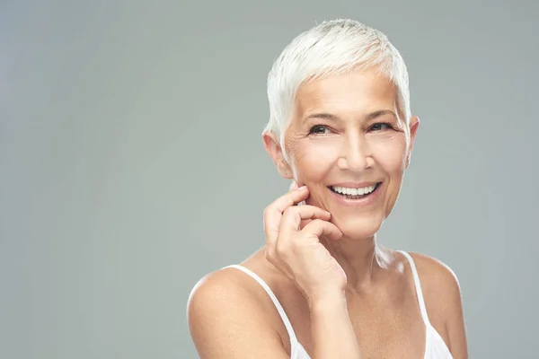 Vackra leende Senior Kvinna med kort grått hår poserar framför grå bakgrund. Skönhet fotografi. — Stockfoto