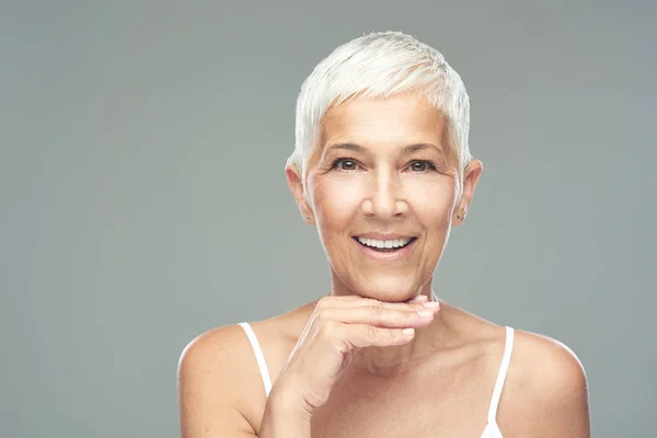 Mooie lachende Senior vrouw met korte grijze haren poseren in de voorkant van de grijze achtergrond. Beauty fotografie. — Stockfoto