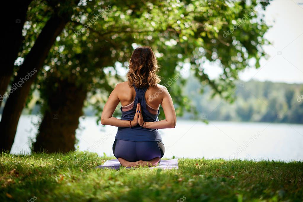 Rear view of Caucasian brunette kneeling on exercise mat and meditating in nature.