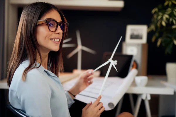 Atractiva mujer de negocios caucásica en traje sentado en la oficina moderna y sosteniendo el modelo de molino de viento. Concepto de desarrollo sostenible . — Foto de Stock