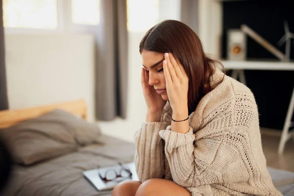 Attractive caucasian brunette dressed in beige sweater sitting on bed in bedroom and having headache. — Stock Photo, Image