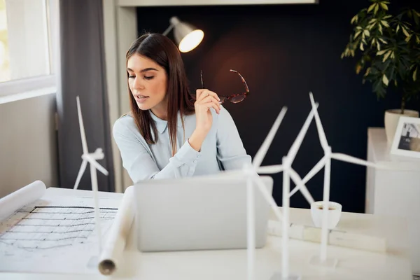 Encantadora morena trabajadora vestida con ropa formal sentada en el escritorio de la oficina, sosteniendo anteojos y mirando planes. En el escritorio son los modelos de ordenador portátil y molino de viento . — Foto de Stock