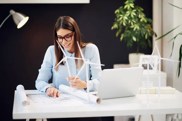 Charmant hardwerkende brunette gekleed in formele slijtage zittend op Bureau in kantoor, het houden van windmolen model en kijken naar plannen. Op de Bureau is een laptop. — Stockfoto