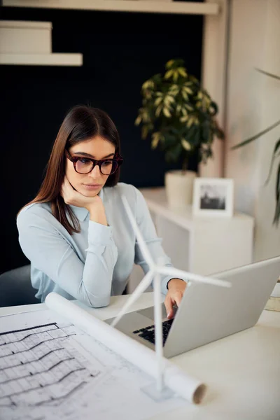 美丽的白人女商人在正式穿着和眼镜坐在办公室，使用笔记本电脑。桌子上有蓝图、笔记本电脑和风车模型。可持续发展理念. — 图库照片