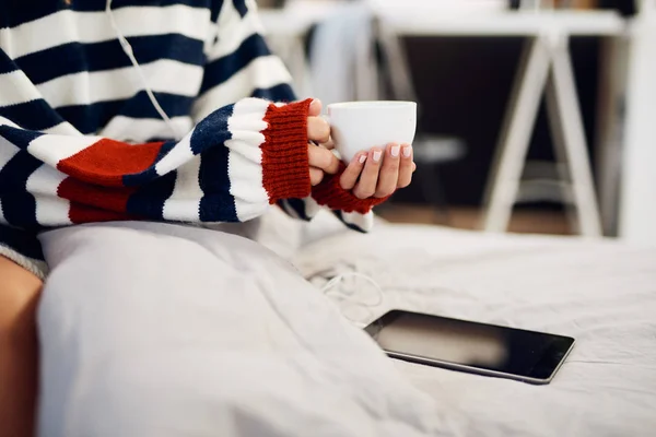 Blanke vrouw in gestreepte trui zittend op bed, luisteren muziek over Tablet en het houden van haar verse koffie in de ochtend. — Stockfoto