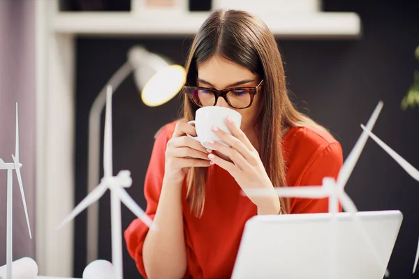 Hermosa mujer de negocios caucásica vestida con blusa roja sentada en la oficina moderna y tomando café. En la mesa son, planos, modelos de ordenador portátil y molino de viento. Concepto de desarrollo sostenible . — Foto de Stock