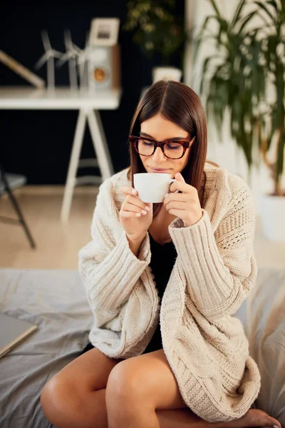 Kaukaski kobieta ubrana w beżowy sweter siedzi na łóżku i trzyma świeżą poranną kawę. — Zdjęcie stockowe