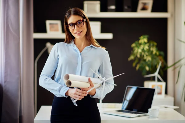 Empresária sorridente atraente no desgaste formal e com óculos em pé no escritório moderno e segurando plantas e modelo de moinho de vento. Conceito de desenvolvimento sustentável . — Fotografia de Stock