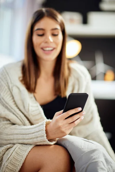 Красивая улыбающаяся кавказская брюнетка сидит в бежевом свитере, сидя на кровати в спальне и используя смартфон. Выборочный фокус на руке . — стоковое фото
