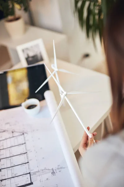 Rear view of caucasian businesswoman holding windmill model and looking at blueprints while sitting in office. Sustainable development concept.