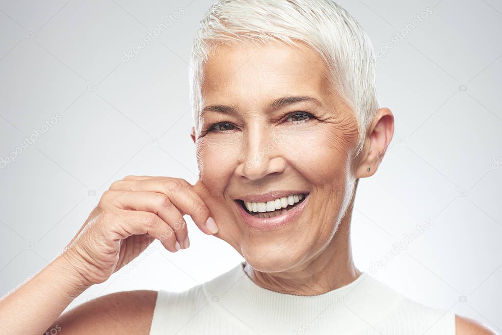 Gorgeous smiling Caucasian senior woman with short gray hair pinching her cheek. Beauty photography.