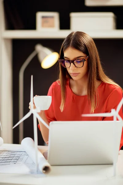 Όμορφη Καυκάσιος επιχειρηματίας ντυμένος με κόκκινη μπλούζα που κάθεται στο μοντέρνο γραφείο και κάνει καφέ. Στο τραπέζι είναι, σχεδιαγράμματα, λάπτοπ και μοντέλα ανεμόμυλος. Έννοια της αειφόρου ανάπτυξης. — Φωτογραφία Αρχείου