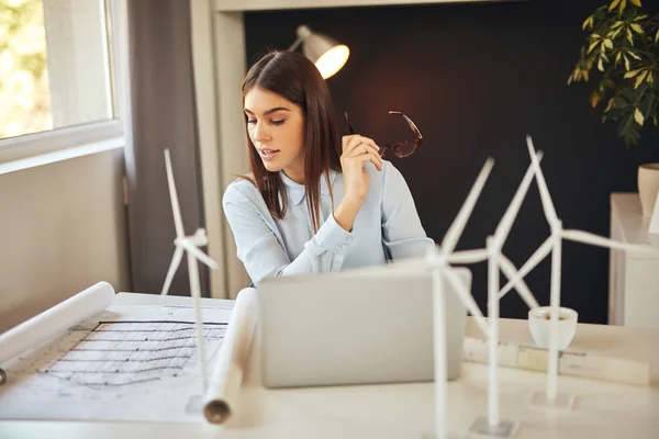 Encantadora morena trabajadora vestida con ropa formal sentada en el escritorio de la oficina, sosteniendo anteojos y mirando planes. En el escritorio son los modelos de ordenador portátil y molino de viento . — Foto de Stock