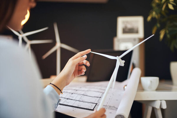 Businesswoman in suit sitting in office and holding windmill model. On desk are coffee, documentation and laptop. Sustainable concept.