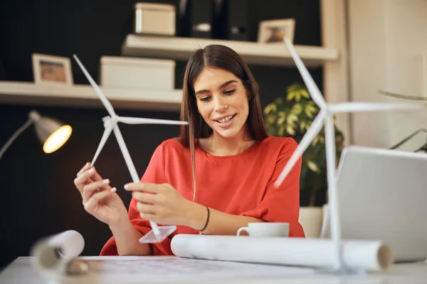 Dedicada mujer de negocios bonita trabajadora vestida con ropa formal sentada en la oficina y sosteniendo el modelo de molino de viento. En la mesa están los planos y el portátil. Concepto de desarrollo sostenible . — Foto de Stock