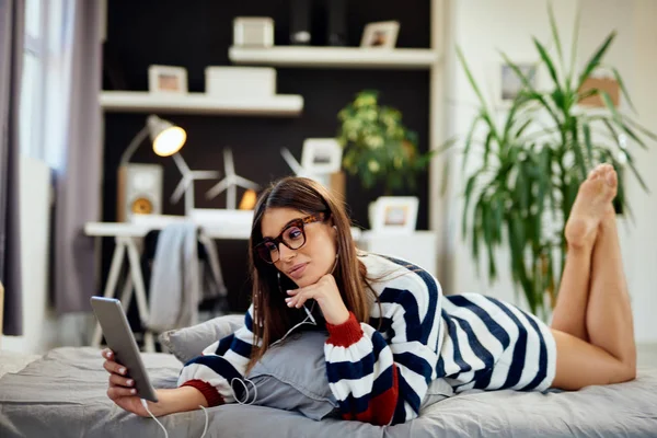 Stunning brunette in sweater lying on stomach on bed in bedroom and using tablet. Morning time. — Stock Photo, Image