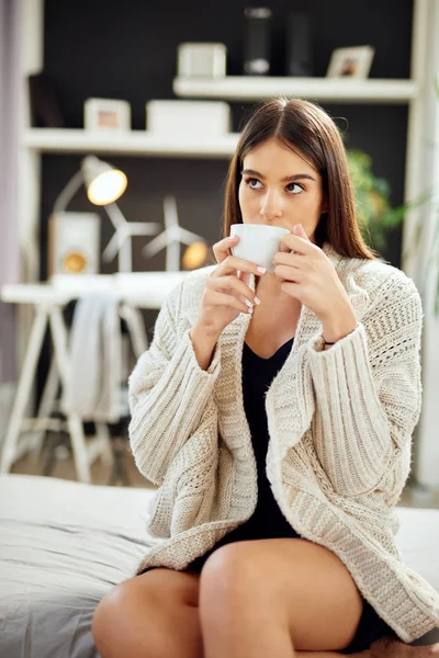 Белая женщина в бежевом свитере сидит на кровати и держит свежий утренний кофе. . — стоковое фото