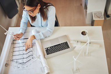 Ofiste masada oturan, yel değirmeni modeli tutan ve planları bakarak resmi giyim giymiş büyüleyici çalışkan esmer üst görünümü. Masanın üzerinde dizüstü bilgisayar.