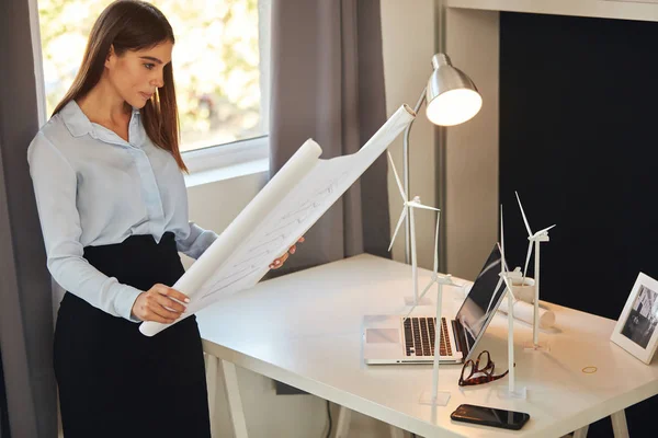 Atractiva mujer de negocios caucásica en camisa y falda de pie en la oficina junto a la ventana y mirando el plano. En el escritorio hay modelos de molino de viento y portátil. Concepto de desarrollo sostenible . — Foto de Stock