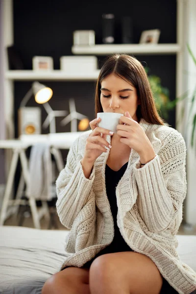 Caucasian woman dressed in beige sweater sitting on bed and holding fresh morning coffee. — Stock Photo, Image