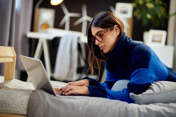 Attraktiv kaukasisk brunett i blå och svart tröja liggande på magen i sängen och skriva på laptop. — Stockfoto