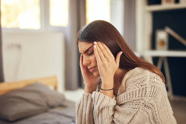 Attractive caucasian brunette dressed in beige sweater sitting on bed in bedroom and having headache. — Stock Photo, Image