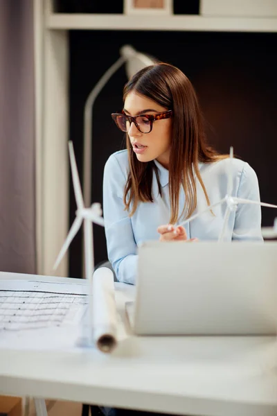 Γοητευτικό εργαζόμενη μελαχρινή ντυμένος με επίσημο ντύσιμο κάθεται στο γραφείο στο γραφείο, κρατώντας το μοντέλο ανεμόμυλος και κοιτάζοντας τα σχέδια. Στο γραφείο είναι φορητός υπολογιστής. — Φωτογραφία Αρχείου
