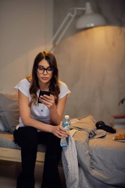 Çekici Kafkas esmer başında kulaklık ile pijama yatakta oturan ve el akıllı telefon. Yatak kahvaltısında. Yatak odası iç.