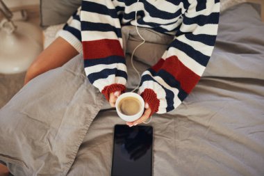 Çizgili kazak giyen beyaz kadın yatakta oturuyor, tablet üzerinden müzik dinliyor ve taze sabah kahvesini tutuyor..