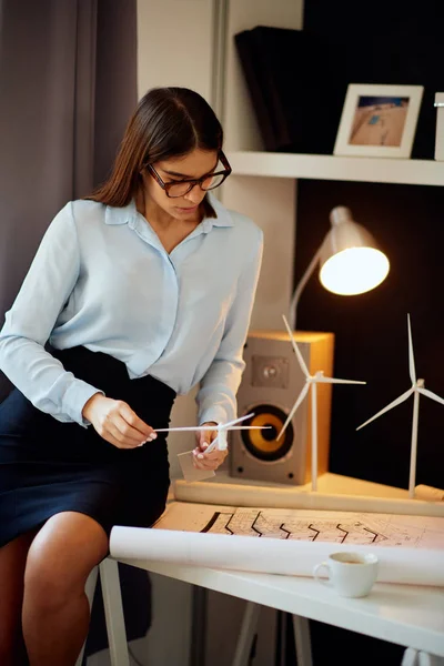Preciosa mujer de negocios caucásica en camisa y falda sentada en el escritorio, sosteniendo modelos de molino de viento y mirando los planes. Concepto de desarrollo sostenible . — Foto de Stock