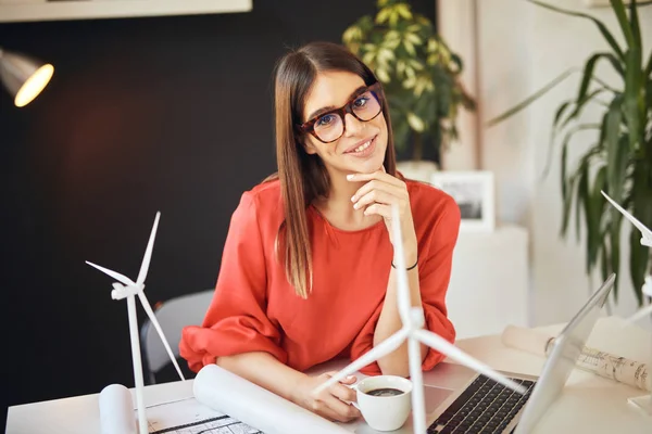 Mooie Kaukasische zakenvrouw gekleed in rode blouse zitten in modern kantoor en driking koffie terwijl het kijken naar de camera. Op tafel liggen blauwdrukken, laptop- en windmolenmodellen. Duurzame ontwikkeling. — Stockfoto