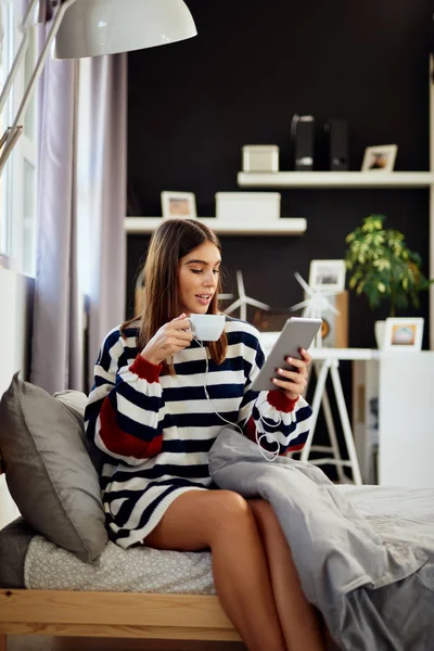 Schöne lächelnde kaukasische Brünette in Pullover auf dem Bett im Schlafzimmer sitzend, mit Tablette und Kaffee trinken in der Früh. — Stockfoto