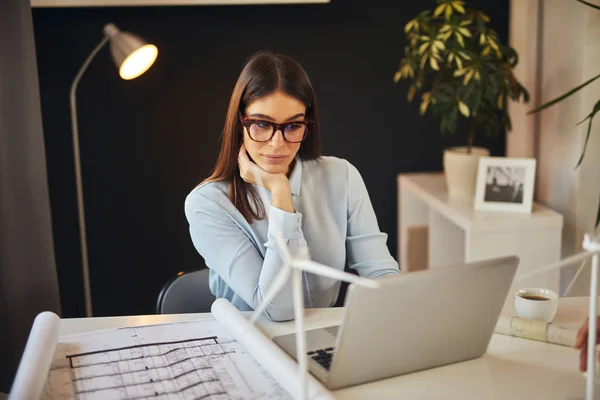 美丽的白人女商人在正式穿着和眼镜坐在办公室，使用笔记本电脑。桌子上有蓝图、笔记本电脑和风车模型。可持续发展理念. — 图库照片