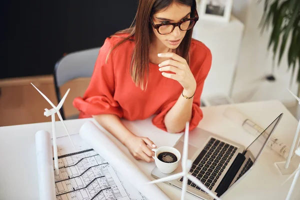 Красивая кавказская деловая женщина в красной блузке сидит в современном офисе, смотрит на ноутбук и высушивает кофе. На столе находятся чертежи, ноутбуки и модели ветряных мельниц. Концепция устойчивого развития . — стоковое фото