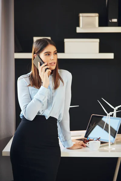 Atractiva mujer de negocios vestida con ropa formal de pie en la oficina, utilizando el teléfono inteligente. En la mesa están los modelos de molino de viento y portátil. Concepto de desarrollo sostenible . — Foto de Stock
