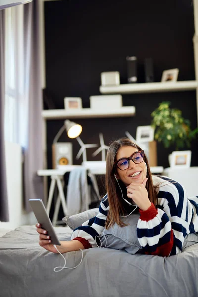 Bedövning brunett i tröja som ligger på magen på sängen i sovrummet och använda Tablet PC. Morgon tid. — Stockfoto