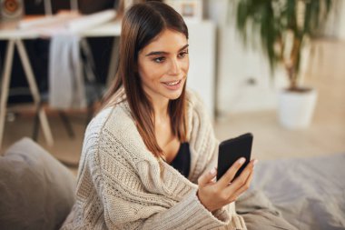 Beyaz tenli, bej kazak giymiş çekici bir esmer yatak odasında oturuyor ve akıllı telefon kullanıyor..