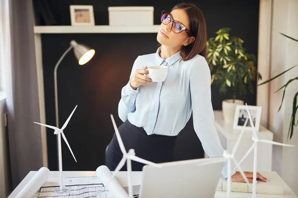 Preciosa mujer de negocios de pie en la oficina y apoyada en el escritorio mientras sostiene la taza de café. En el escritorio son, planes, modelos de molino de viento y portátil. Concepto de desarrollo sostenible . — Foto de Stock