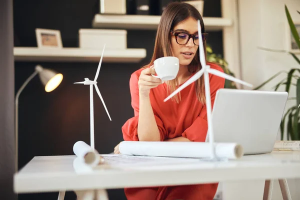 Hermosa mujer de negocios caucásica vestida con blusa roja sentada en la oficina moderna y tomando café. En la mesa son, planos, modelos de ordenador portátil y molino de viento. Concepto de desarrollo sostenible . — Foto de Stock