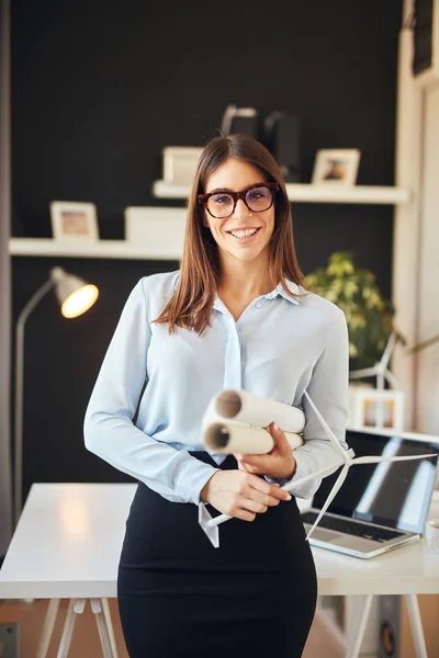 Aantrekkelijke lachende zakenvrouw in formele slijtage en met brillen staande in moderne kantoor en het houden van blauwdrukken en windmolen model. Concept duurzame ontwikkeling. — Stockfoto