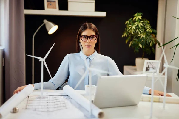 Привлекательная кавказская деловая женщина в рубашке и юбке и в очках, позирующая в офисе. На столе находятся ноутбуки, планы и модели ветряной мельницы. Концепция устойчивого развития . — стоковое фото