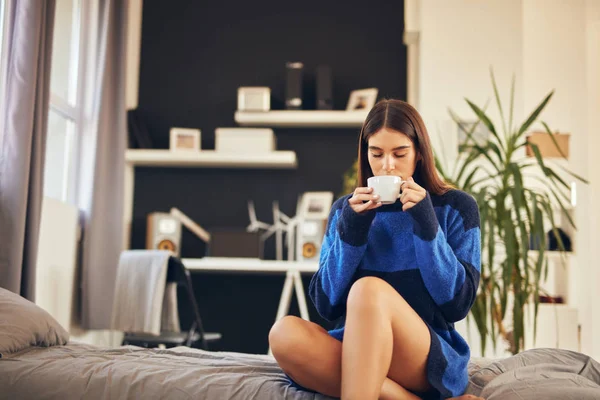 Charmante brunette in blauwe trui zittend in de slaapkamer in de ochtend en koffie drinken. — Stockfoto