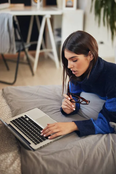Atraktivní běloška brunetka v modrém a černém svetru ležící na břiše v posteli a psaní na notebooku. — Stock fotografie