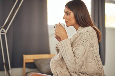 Bej kazak giymiş beyaz kadın yatakta oturan ve taze sabah kahvesi tutan.