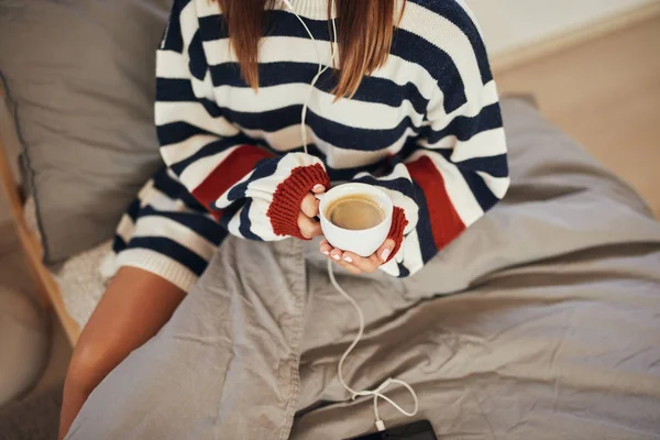 Kaukaski kobieta w paski sweter siedzi na łóżku, słuchanie muzyki nad tabletem i trzymając jej świeżą poranną kawę. — Zdjęcie stockowe