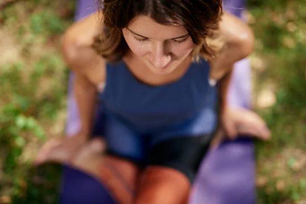 Vista dall'alto della bella bruna cucasiatica vestita con abbigliamento sportivo seduta su un tappeto in natura in posa yoga al ginocchio . — Foto Stock