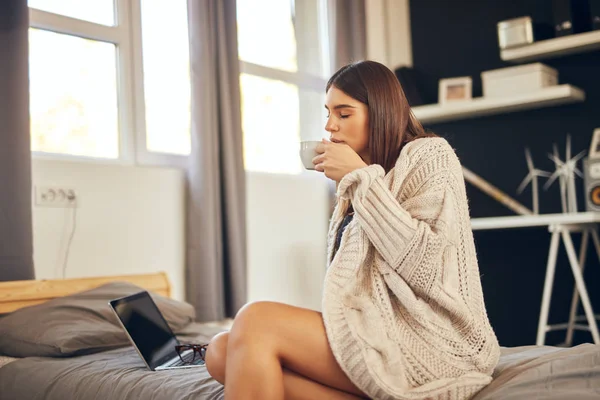 Caucasian woman dressed in beige sweater sitting on bed and holding fresh morning coffee. — Stock Photo, Image